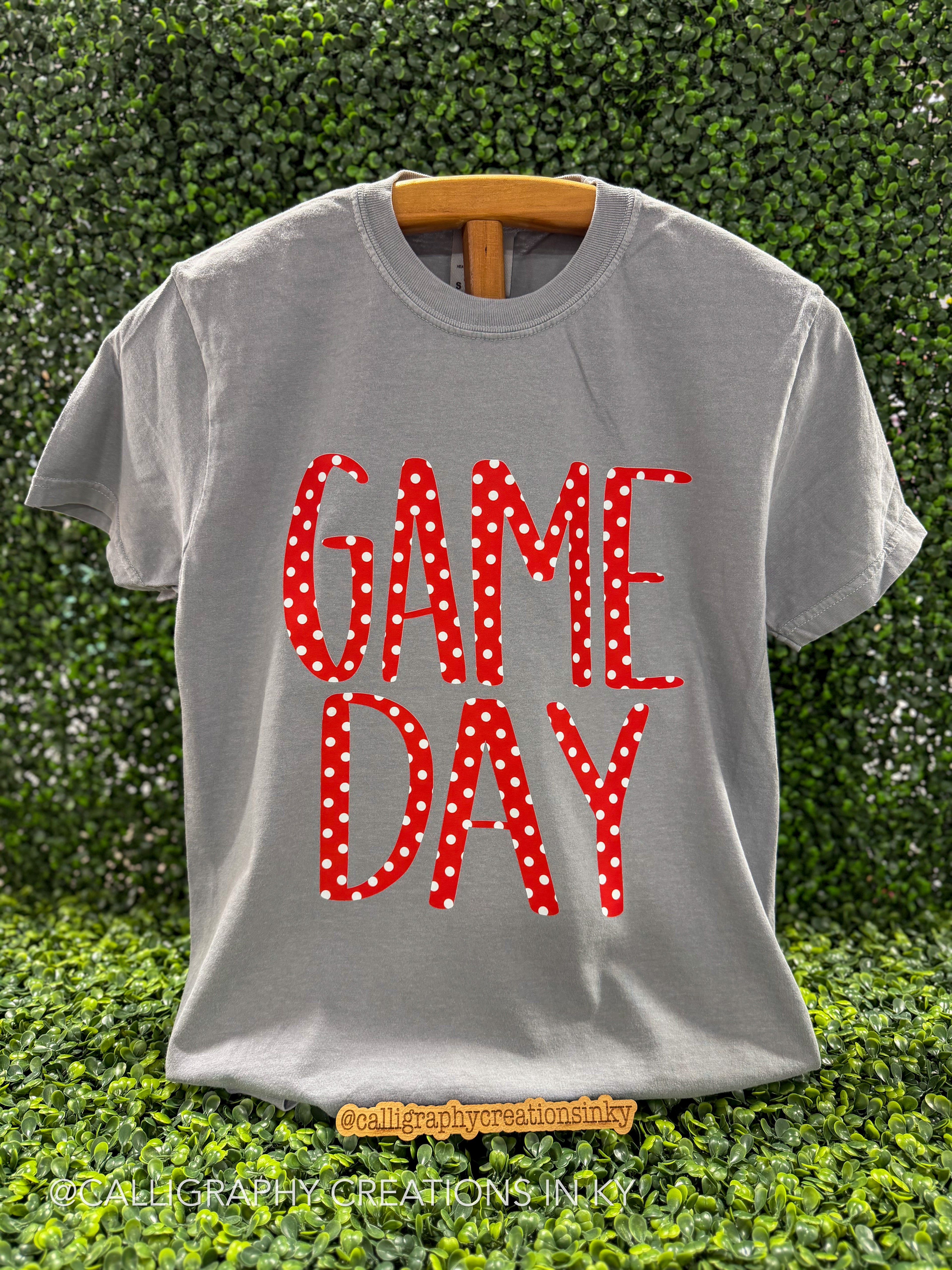 A gray short-sleeve t-shirt on a wooden hanger, featuring the words 'GAME DAY' in red letters with white polka dots, set against a green hedge background.