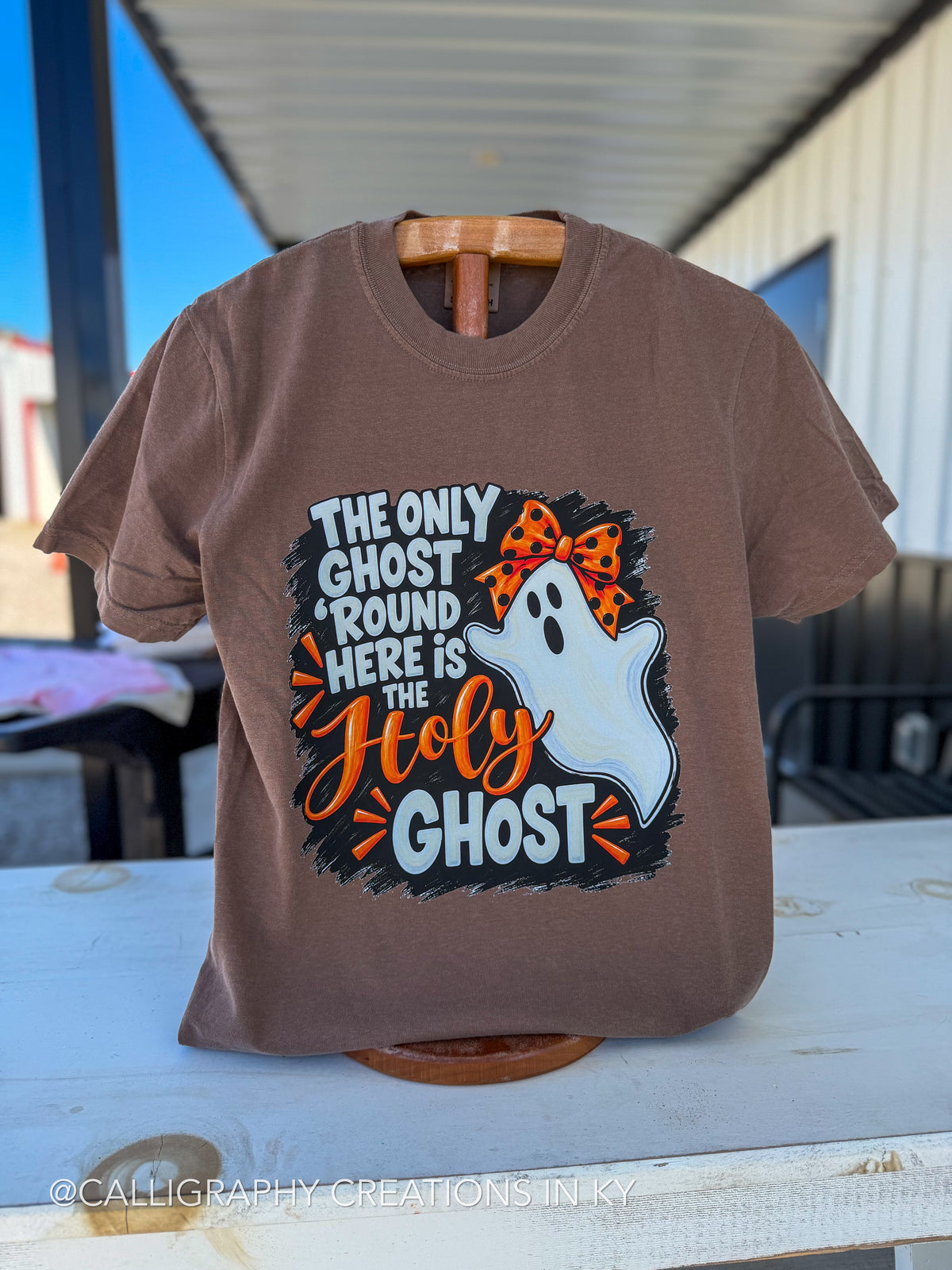 A brown short-sleeve graphic t-shirt displayed on a wooden hanger on an outdoor porch table. The shirt features a Halloween graphic with the text 'THE ONLY GHOST ROUND HERE IS THE Holy GHOST' in white and orange lettering, alongside a white ghost wearing an orange and black leopard print bow.