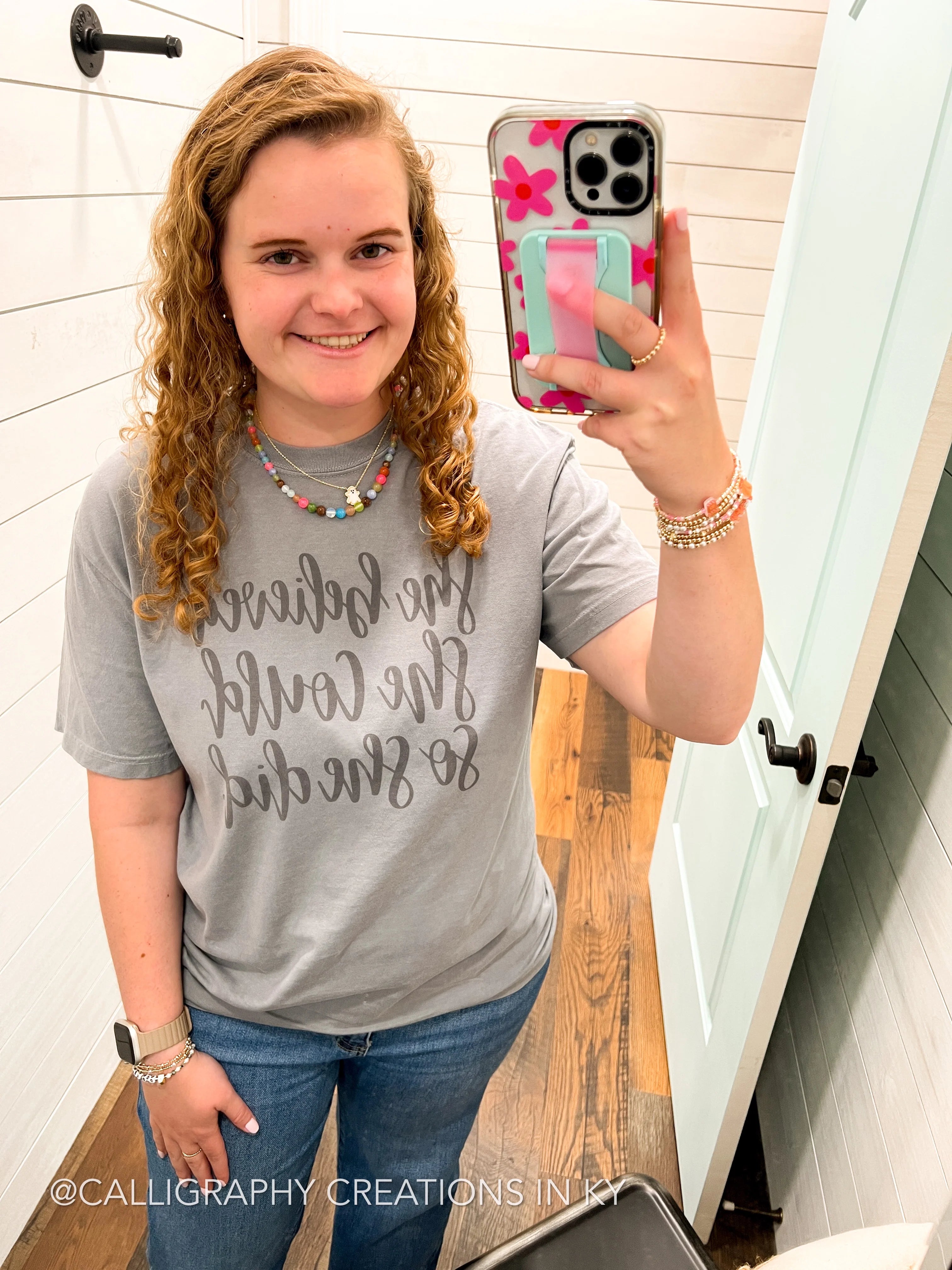 Person taking a mirror selfie wearing a gray t-shirt with text and blue jeans.