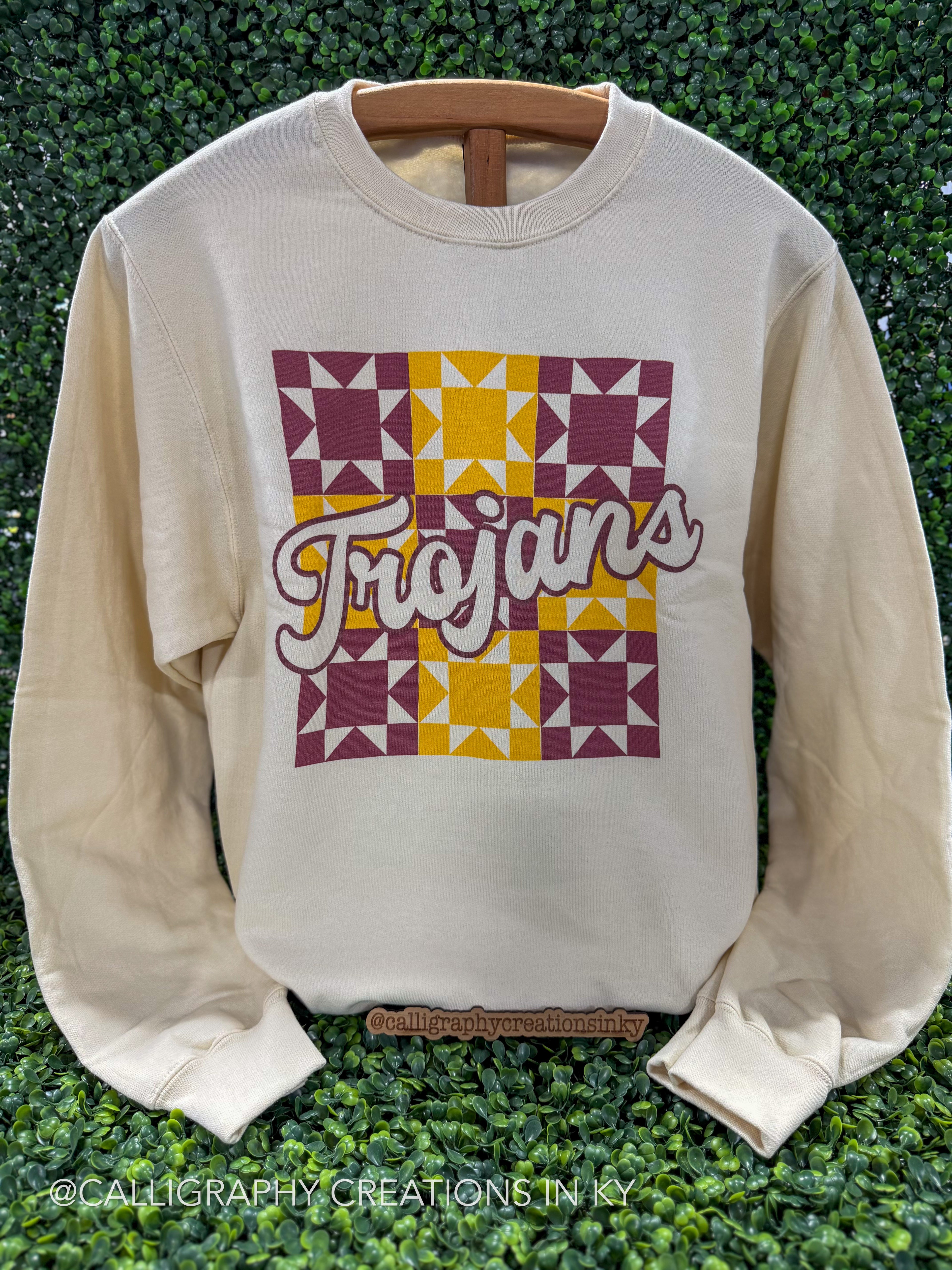An off-white long-sleeve crewneck sweatshirt with a graphic on the front. The graphic features a square quilted pattern in maroon and yellow, with the word 'Trojan' written across it in a white cursive font with a maroon outline.
